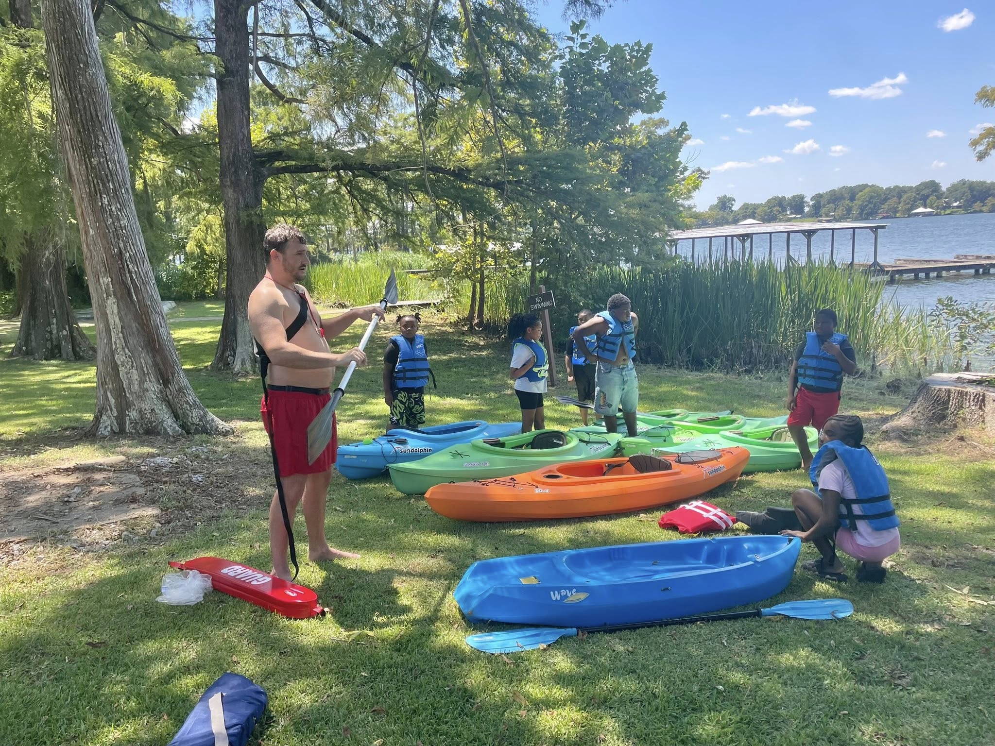 A man and a group of kids standing by some kayaks on the ground.